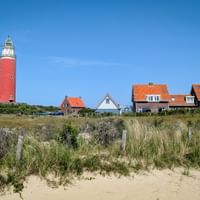 Red lighthouse on Texel island with traditional Dutch houses with orange roofs, surrounded by dune grass and sandy beach under blue sky.