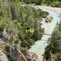 Türkisfarbene Isar fließt durch dichten Wald mit felsigen Ufern. Eine kleine Fußgängerbrücke überquert den Fluss in der Münchner Seenregion.