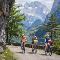 Drei Radfahrer fahren am Gosausee entlang mit dramatischen Dachstein-Gipfeln im Hintergrund. Felswand links, Alpensee und Wald sichtbar.