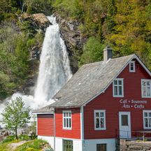 Im Vordergrund ein landestypisches rotes Holzhaus, im Hintergrund der Steinsdals Wasserfall