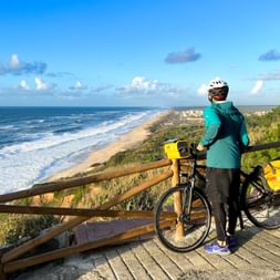 Cyclist in turquoise jacket with helmet standing at wooden viewpoint overlooking Atlantic beach near Quiaios, Coimbra. Yellow panniers on bicycle.