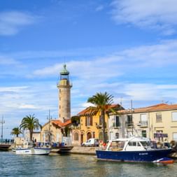 Stone lighthouse and colorful Mediterranean buildings along the harbor in Le Grau-du-Roi, Camargue. Blue fishing boats moored in the water.