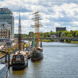Historische Segelschiffe im Bremer Museumshafen an der Weser, mit modernen Gebäuden und einer Brücke im Hintergrund unter bewölktem Himmel.
