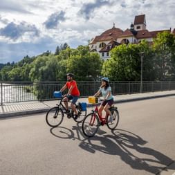 Two cyclists with helmets and panniers riding across a bridge over a river in Füssen. Historic buildings with red roofs and green trees visible.