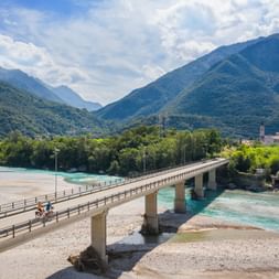 Cyclists crossing a concrete bridge over the turquoise Tagliamento River near Venzone, with forested mountains and a small village in the background.
