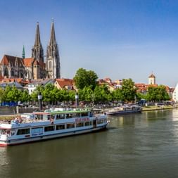 Regensburger Dom mit zwei Türmen an der Donau. Ausflugsschiffe am Ufer mit historischen Gebäuden und Bäumen.