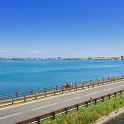 Zwei Radfahrer auf einer Brücke über eine blaue Lagune bei Grado. Die Küstenstadt ist in der Ferne unter blauem Himmel sichtbar.