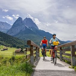 Two cyclists crossing a wooden bridge on a bike path near Garmisch, Bavaria, with dramatic mountain peaks and green meadows in the background.