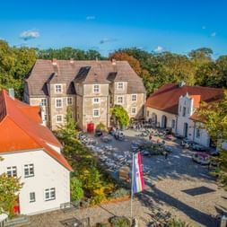 Luftaufnahme des Wasserschlosses Mellenthin mit Steingebäuden, roten Ziegeldächern und Besuchern in Außenbereichen unter blauem Himmel.