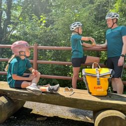 Family of three in matching teal cycling gear taking a break on a wooden bench. Child in pink helmet sits on bench, parents stand behind. Yellow Eurobike bag visible.