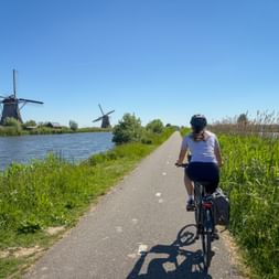Cyclist with panniers riding on paved path along canal in Kinderdijk. Historic windmills visible in background under clear blue sky.