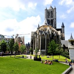Nikolauskirche in Gent mit gotischem Turm und Türmchen. Menschen entspannen auf der grünen Wiese vor der mittelalterlichen Kirche.