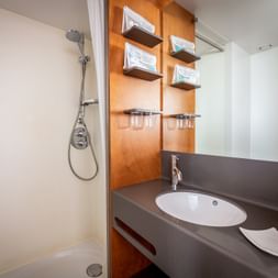Compact bathroom on MS Ave Maria with shower, sink, mirror, wooden shelves with towels, and gray countertop.