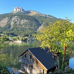 Hölzernes Bootshaus am Altausseer See mit bewaldetem Berg und felsiger Spitze unter klarem blauen Himmel im Salzkammergut, Österreich.