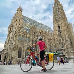 Radfahrerin mit rotem Fahrrad und gelben Taschen steht vor dem Stephansdom in Wien und zeigt auf das gotische Gebäude.
