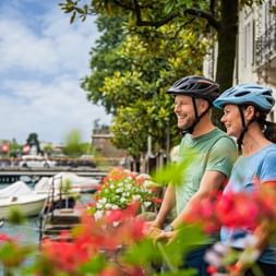 Two smiling cyclists with helmets standing by a flower-lined canal in Peschiera, Lake Garda, with boats and historic buildings in the background.