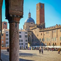 Blick auf die Piazza Sordello in Mantua durch Steinsäulen, mit Kopfsteinpflasterplatz, Domkuppel, hohem Backsteinturm und historischen Gebäuden.