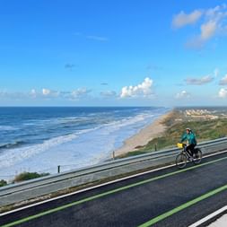 Radfahrer auf Radweg entlang der Atlantikküste bei Quiaios, Portugal. Langer Sandstrand erstreckt sich in die Ferne unter blauem Himmel.
