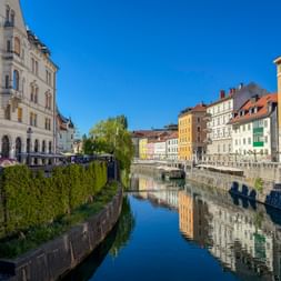 Colorful historic buildings lining the Ljubljanica River in Ljubljana, Slovenia. The calm water reflects the architecture under a clear blue sky.