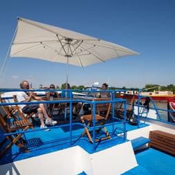 Sonnendeck der MS Caprice mit blauem Boden, weißem Sonnenschirm, Holzliegestühlen und entspannenden Gästen. Fluss und Boote im Hintergrund.