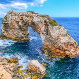 Es Pontàs natürlicher Felsbogen erhebt sich aus türkisfarbenem Mittelmeer vor Mallorcas Küste unter blauem Himmel mit weißen Wolken.