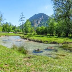 Clear river Ammer flowing through green meadows with trees and a forested mountain peak under blue sky in Bavaria, Germany.