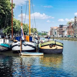Traditionelle Segelboote im Hafen von Dordrecht mit historischen niederländischen Gebäuden entlang der Uferpromenade unter blauem Himmel.