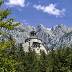 Hohenwerfen Castle with green copper dome surrounded by forest, with dramatic Tennengebirge mountain peaks rising behind under blue sky.