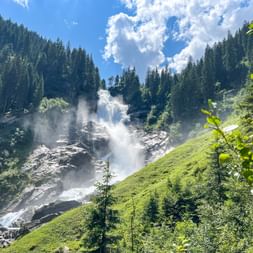 Upper Krimml Waterfall cascading down rocky slopes surrounded by green meadows and dense forest. White mist rises from the falls under blue sky.