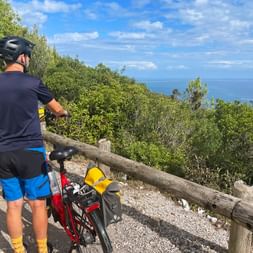 Radfahrer in blauer Hose mit rotem Fahrrad an Holzgeländer, mit Blick auf blaues Meer und grüne Vegetation an Kretas Küste.