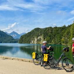 Two cyclists with helmets and panniers pause at the shore of a turquoise alpine lake, surrounded by forested mountains under blue sky.