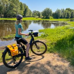Radfahrerin mit Helm und orangen Packtaschen steht neben ihrem Rad am Canal du Midi mit Blick auf ruhiges Wasser und grüne Landschaft.