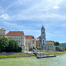 Stift Dürnstein monastery with blue baroque tower on the Danube riverbank in Wachau. Historic buildings line the waterfront with green hills behind.