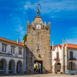 Stone clock tower Torre do Relógio in Caminha's town square, flanked by white buildings with arched facades and orange tile roofs.