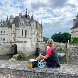 Radfahrerin sitzt auf Steinmauer mit Blick auf Schloss Chenonceau mit Bogenbrücke über dem Fluss Cher unter bewölktem Himmel.
