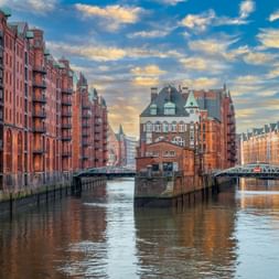 Kanal in der Hamburger Speicherstadt mit roten Backsteinspeichern auf beiden Seiten, zwei Brücken und historischen Gebäuden unter dramatischem Himmel.