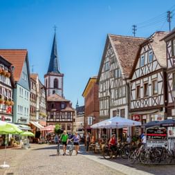 Historischer Marktplatz in Lohr am Main mit bunten Fachwerkhäusern, Kirchturm, Straßencafés und Radfahrern auf Kopfsteinpflaster.