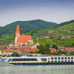 MS Manon Flusskreuzfahrtschiff auf der Donau vor Weißenkirchen mit Kirche mit rotem Dach und terrassierten Weinbergen in der Wachau.
