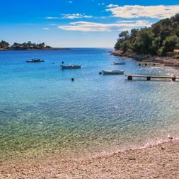 Pebble beach near Pula with clear turquoise water, small boats, and a wooden pier. Green trees line the shore under a blue sky.