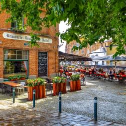 Kopfsteinplatz in Brügge mit Backstein-Brasserie und Café-Terrasse unter Bäumen. Gäste sitzen an Tischen mit roten Stühlen unter Sonnenschirmen.