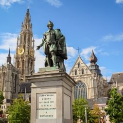 Bronze statue of Peter Paul Rubens on stone pedestal with Cathedral of Our Lady's Gothic tower in background under blue sky in Antwerp.