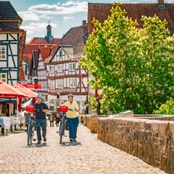 Two cyclists walking their bikes through Melsungen's cobblestone square, surrounded by traditional half-timbered houses and a café terrace.