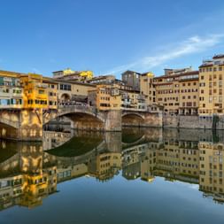 Ponte Vecchio Brücke mit mittelalterlichen Gebäuden über dem Arno in Florenz. Perfekte Spiegelung im ruhigen Wasser unter blauem Himmel.