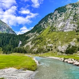 Turquoise Krimml Ache river flowing through green meadow in mountain valley with forested slopes and rocky peaks under blue sky.
