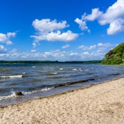 Sandy beach at Roskilde Fjord with gentle waves, forested shores, and blue sky with white clouds in Denmark.