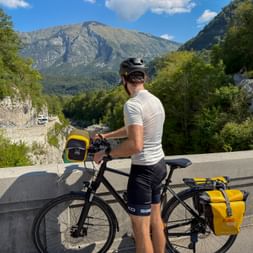 Cyclist with yellow panniers on touring bike pausing on mountain road near Kobarid, Slovenia, with dramatic mountain peaks in background.