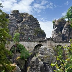 Historic Bastei Bridge with stone arches spanning between towering sandstone rock formations, surrounded by green trees under blue sky.