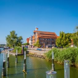 Historisches Fachwerkhaus mit rotem Ziegeldach am Hafen Wolgast. Holzpfähle säumen das grüne Ufer mit Schilf und Bäumen unter blauem Himmel.