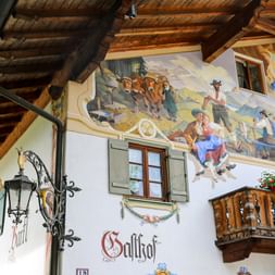 White Bavarian house in Garmisch with colorful Lüftlmalerei frescoes depicting rural scenes. Wooden balcony, shuttered windows, and ornate lantern visible.