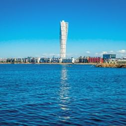 Malmö waterfront with the distinctive twisted Turning Torso skyscraper dominating the skyline. Blue water in foreground, residential buildings along shore.